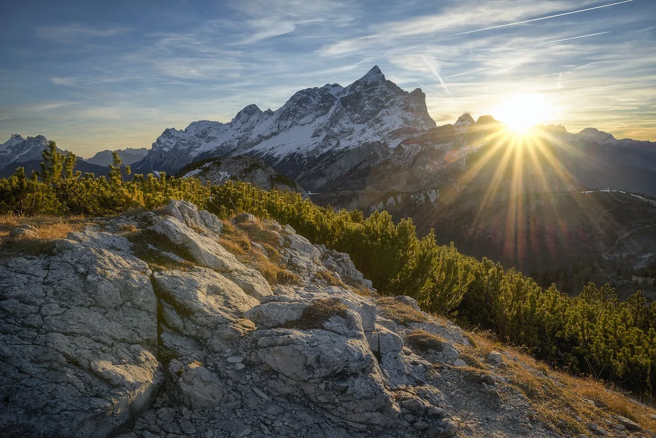 Sun rising beside snow-covered mountains above a rocky slope